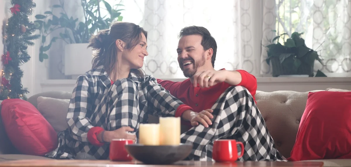 Couple sitting on a couch laughing in matching buffalo plaid pajamas with red mugs and Christmas greenery hung in the background
